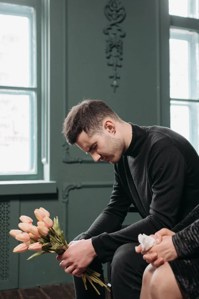 A young man in black holds flowers, symbolizing loss and mourning, seated indoors.